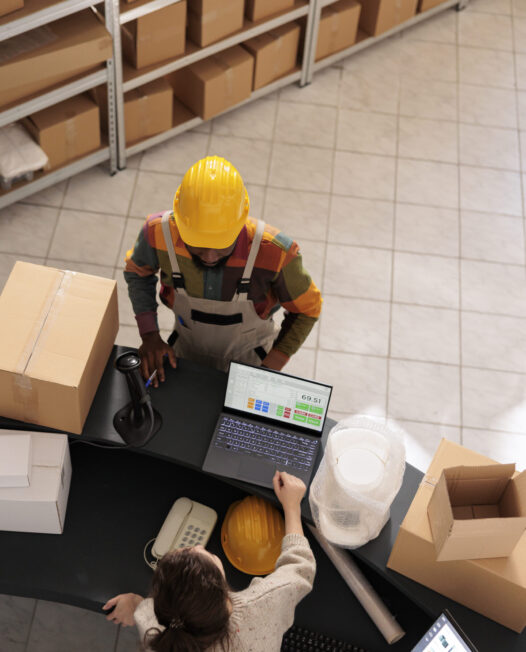 Top view of diverse team discussing customer order preparing packages for delivery in warehouse. African american employee wearing industrial overall and helmet working at goods quality control